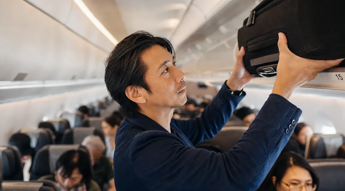 Traveler hoisting a packed carry-on roller bag into an overhead airplane bin