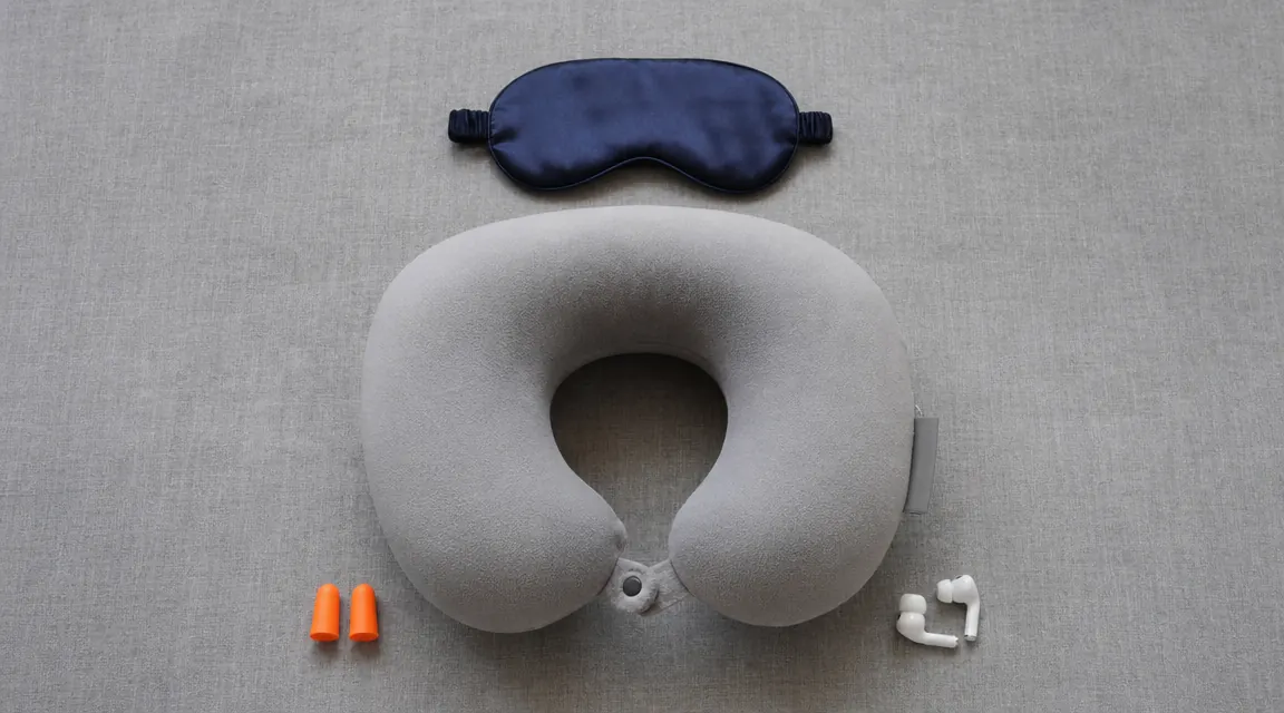 Overhead flat-lay of economy-seat sleep kit: neck pillow, sleep mask, ear plugs, and a pair of noise-canceling earbuds on a grey linen surface